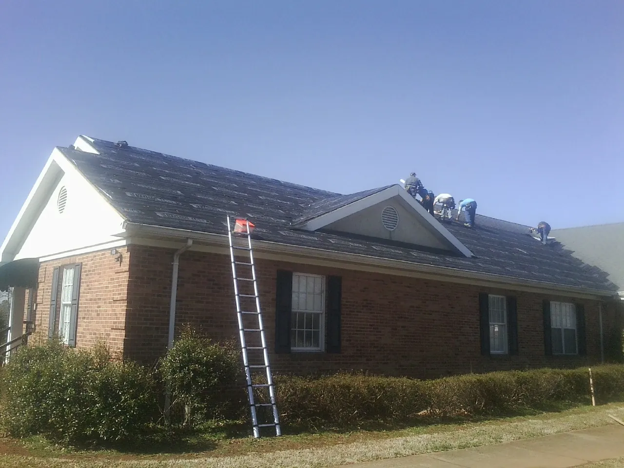 Crew tearing shingles off a brick ranch roof with a ladder set — Metal Roof Repair work by our crew in Belford